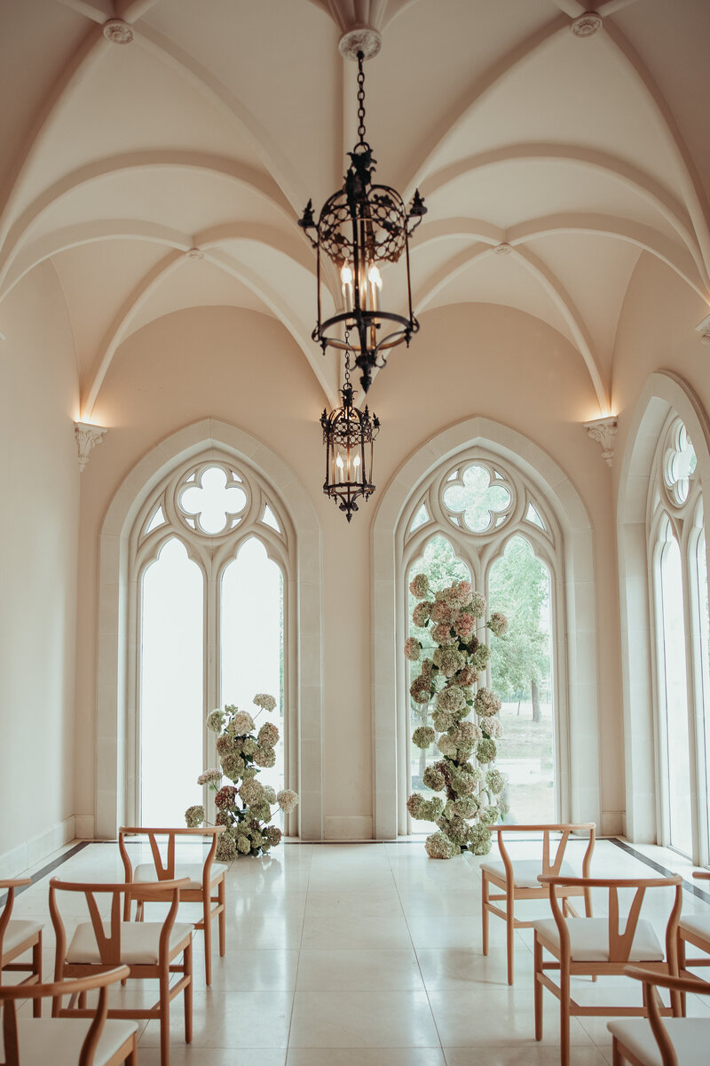 Light-filled ceremony with gothic arched windows, hanging lanterns, sculptural hydrangea pillars, and wishbone chairs.