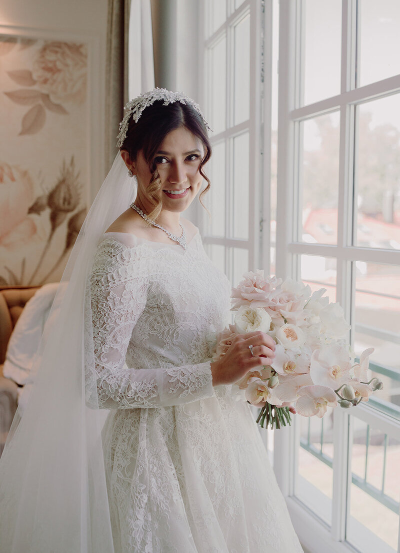 Bride in Elie Saab wedding dress standing by a window before the wedding ceremony, holding a bouquet of pale roses.