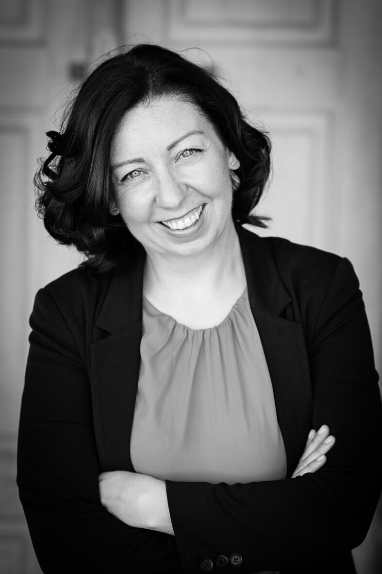 A woman with shoulder-length dark hair, wearing a blazer over a blouse, smiles with arms crossed in a black and white photo, embodying the confidence of an academic leader in post-secondary education.