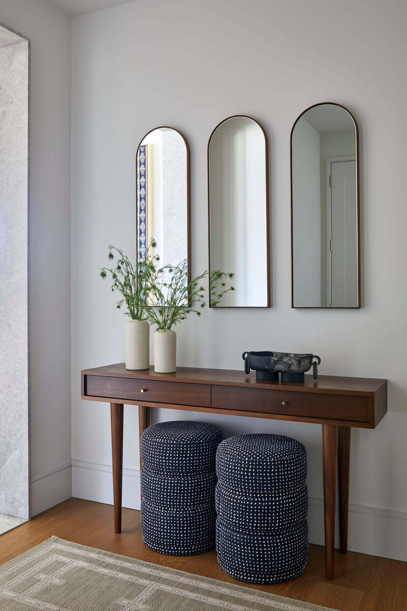 Natural lighting entryway with unique wooden console tables and textured cushion stools. Three arched mirrors on display on wall.