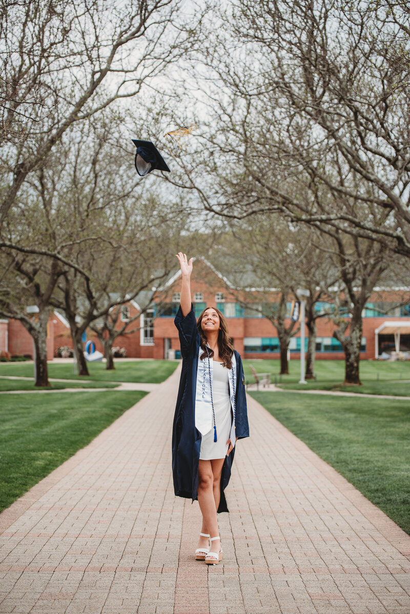 A joyful college graduate in a navy cap and gown throws her graduation cap into the air while walking down a tree-lined campus pathway. Captured during a Connecticut graduation photography session by Nicole Mele Photography, celebrating achievement and new beginnings.