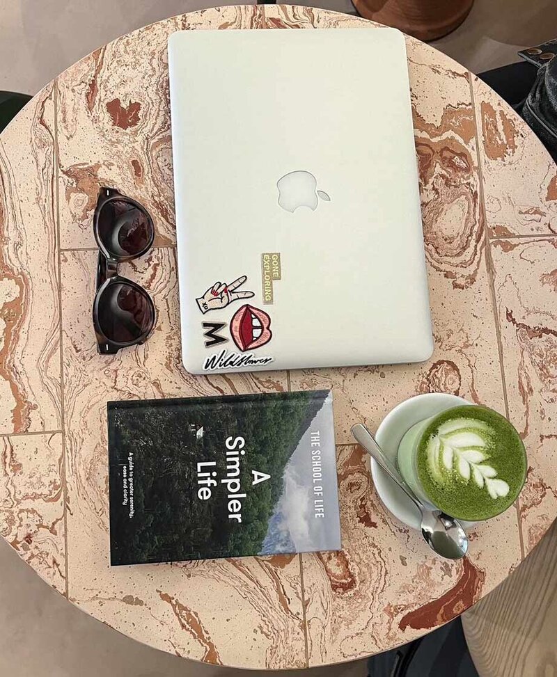 Table at a cafe with cup of matcha, laptop, sunglasses, and book.