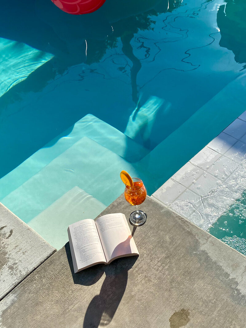 Glass of cocktail next to an open book by a pool