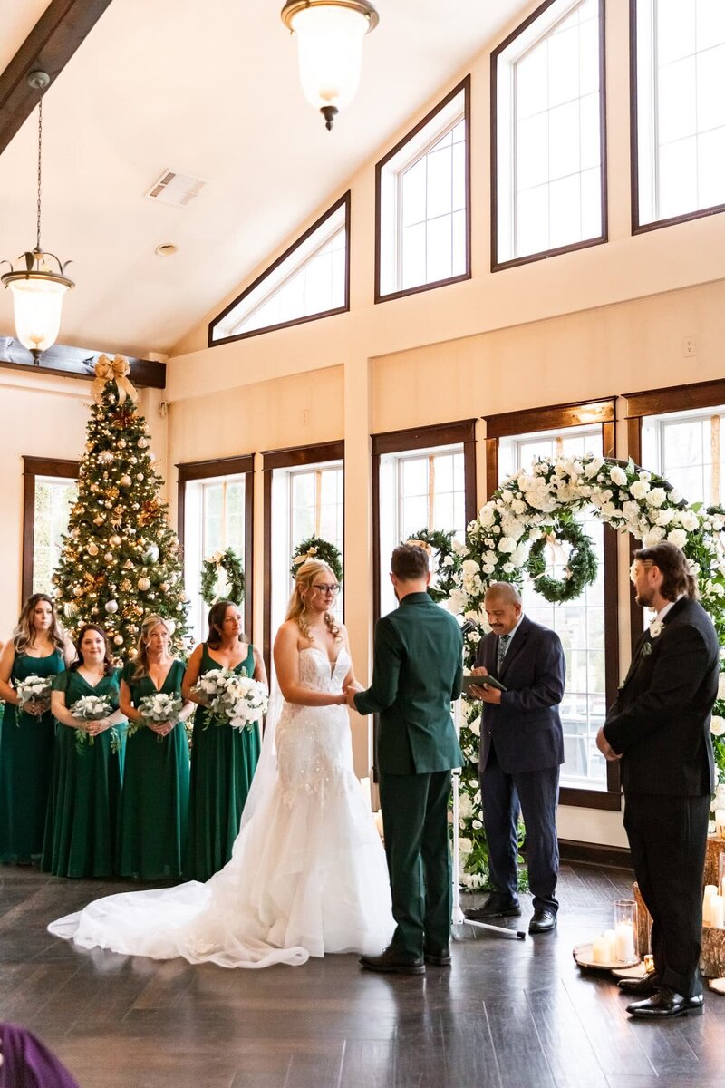 The bride and groom stand hand in hand before an officiant, exchanging vows beneath a lush floral and greenery arch. Bridesmaids in deep emerald dresses clutch bouquets of white roses, standing beside a sparkling Christmas tree as natural light pours through the venue’s floor-to-ceiling windows