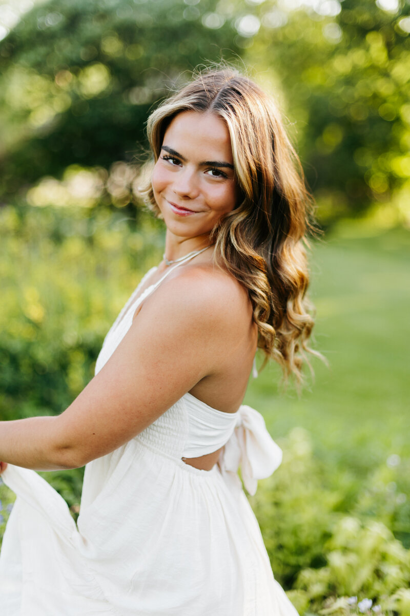 Senior girl in a white dress posing in front of green grass and trees