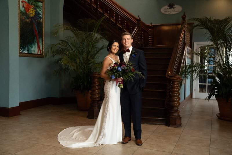 A bride in an elegant white lace gown with train and groom in navy suit pose together holding a colorful bouquet in front of an ornate wooden staircase with tropical plants at The White Room venue in historic downtown St. Augustine