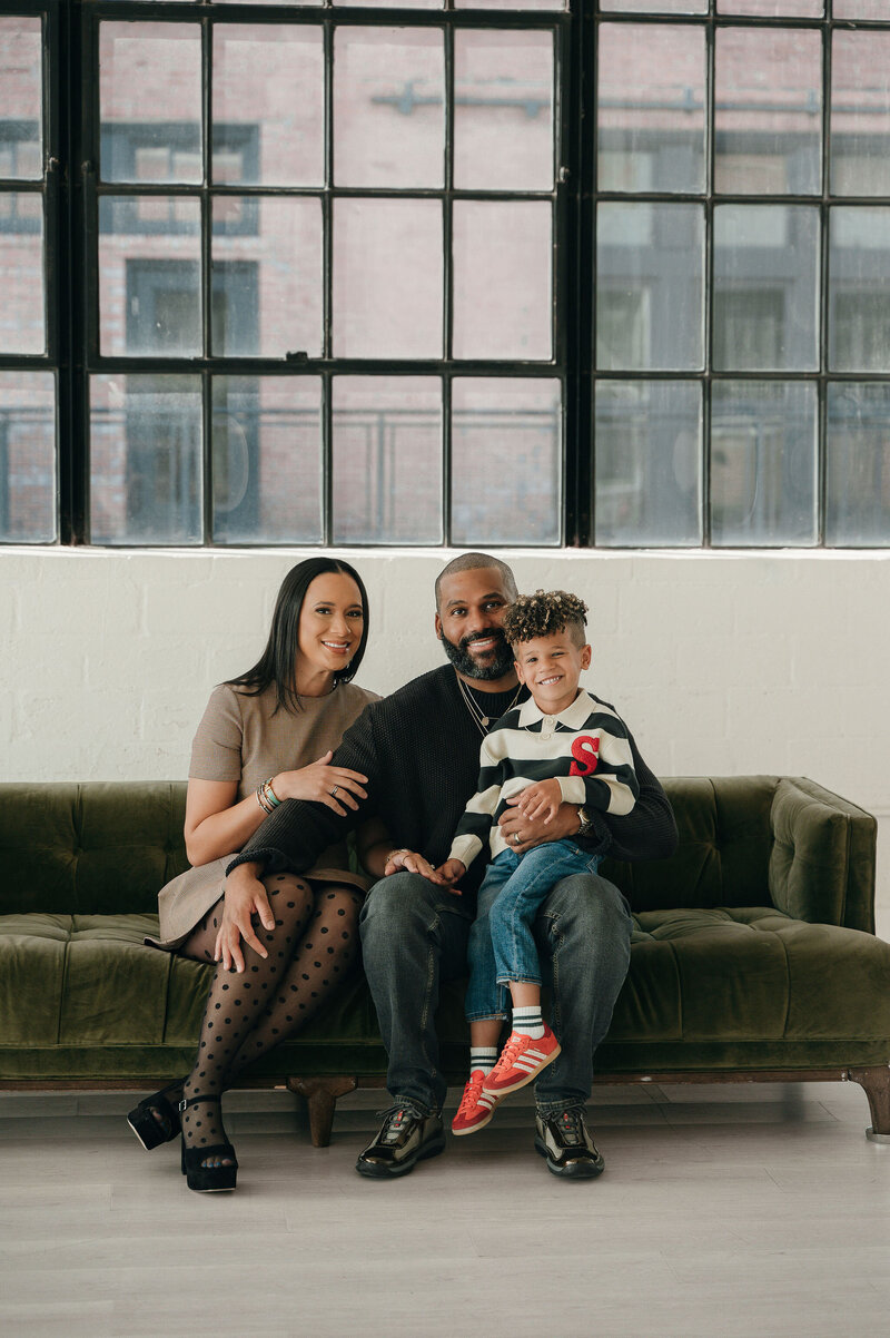 A smiling family sits on a green sofa in front of snowy Christmas trees. The parents hold a baby wearing a headband. The setting is festive and warm.