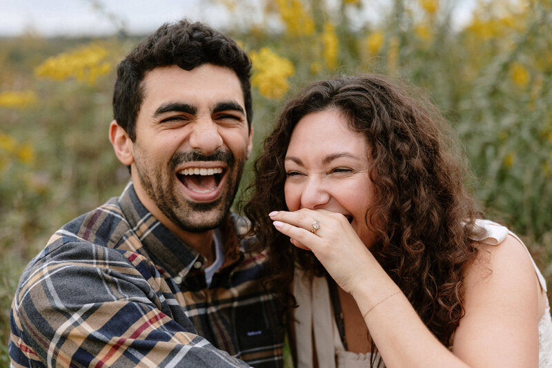  A couple enjoying a lighthearted moment, laughing together in a beautiful field filled with greenery and flowers.