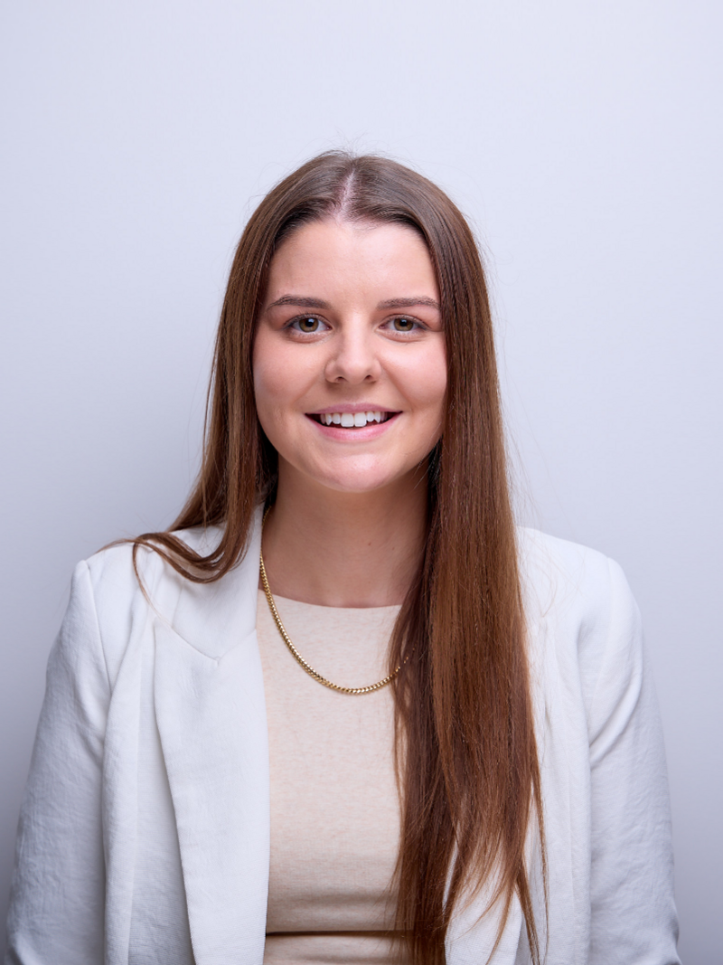 A woman with long hair, wearing a smile. She is standing in front of a wall and her layered hair is visible in the portrait. The focus is on her face, neck, and lips.