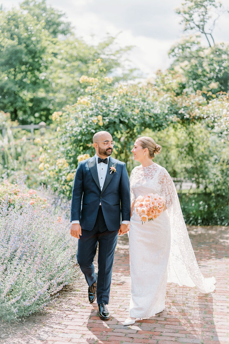 Newport Wedding Photographer | A bride in a white gown and groom in a navy suit walk hand in hand on a sunny day in a lush garden, surrounded by greenery and flowers.