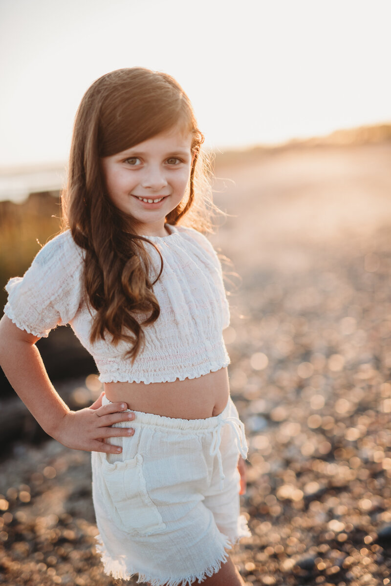 A young girl in a white summer outfit smiles confidently on a sunlit beach during golden hour. Captured during a children’s portrait session in Connecticut by Nicole Mele Photography, highlighting natural light and joy.