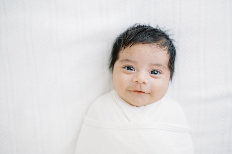 Newborn baby boy face with dark hair and dark eyes looks and smiles wrapped in a white swaddle by Portland Newborn Photographer Emilie Phillipson Photography.