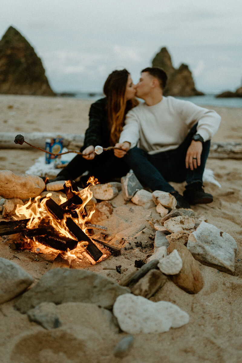 B & T sitting in front of a campfire on the beach, roasting marshmellows for smores while kissing