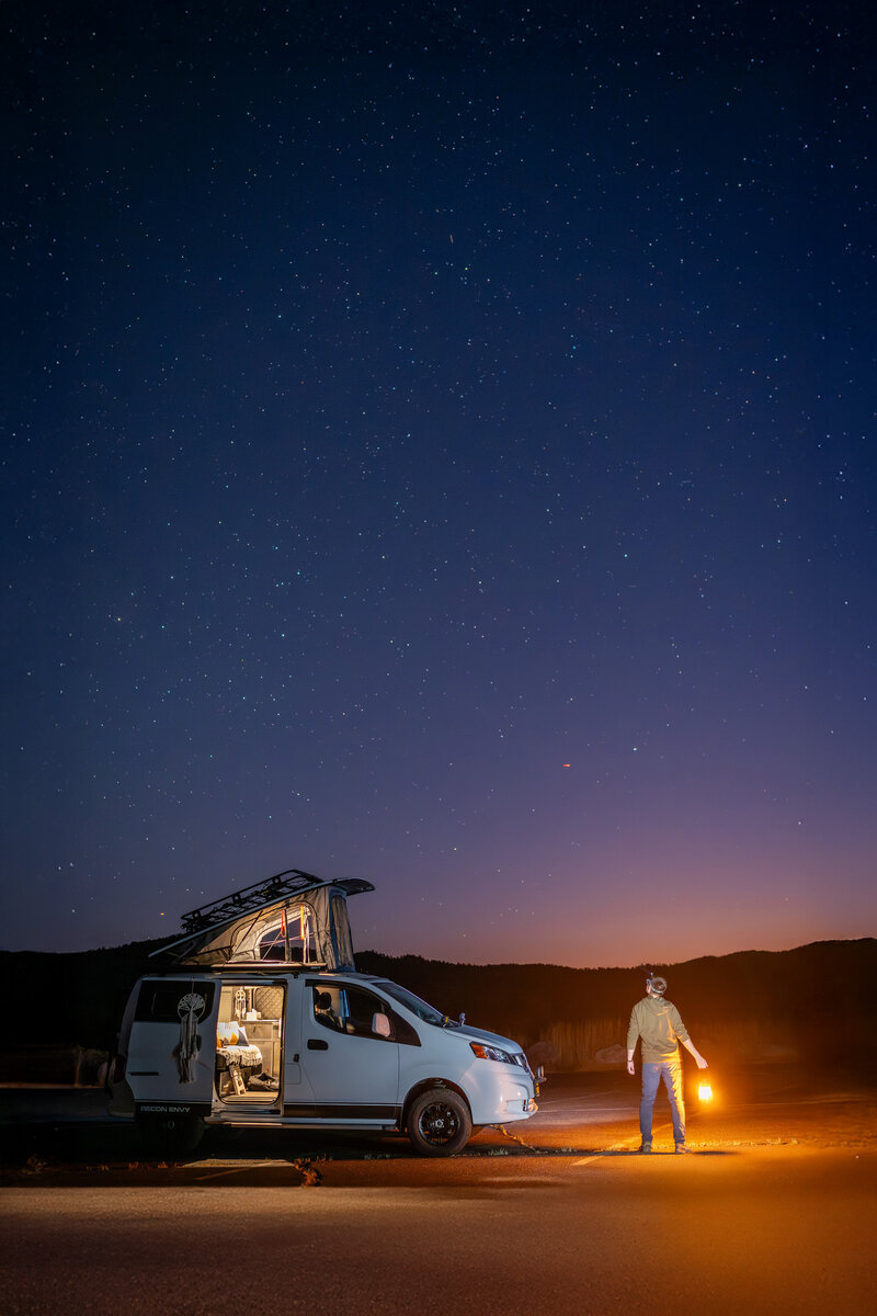 California Elopement Photographer standing beside a camper van in Joshua Tree at night, holding a lantern under starry skies — symbolizing adventure elopement vibes and off-the-beaten-path storytelling.