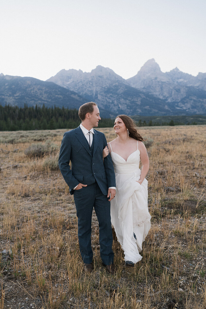Bride and groom standing chest to chest with the brides hands wrapped behind the grooms neck in Grand Teton National Park looking behind them towards a field. 