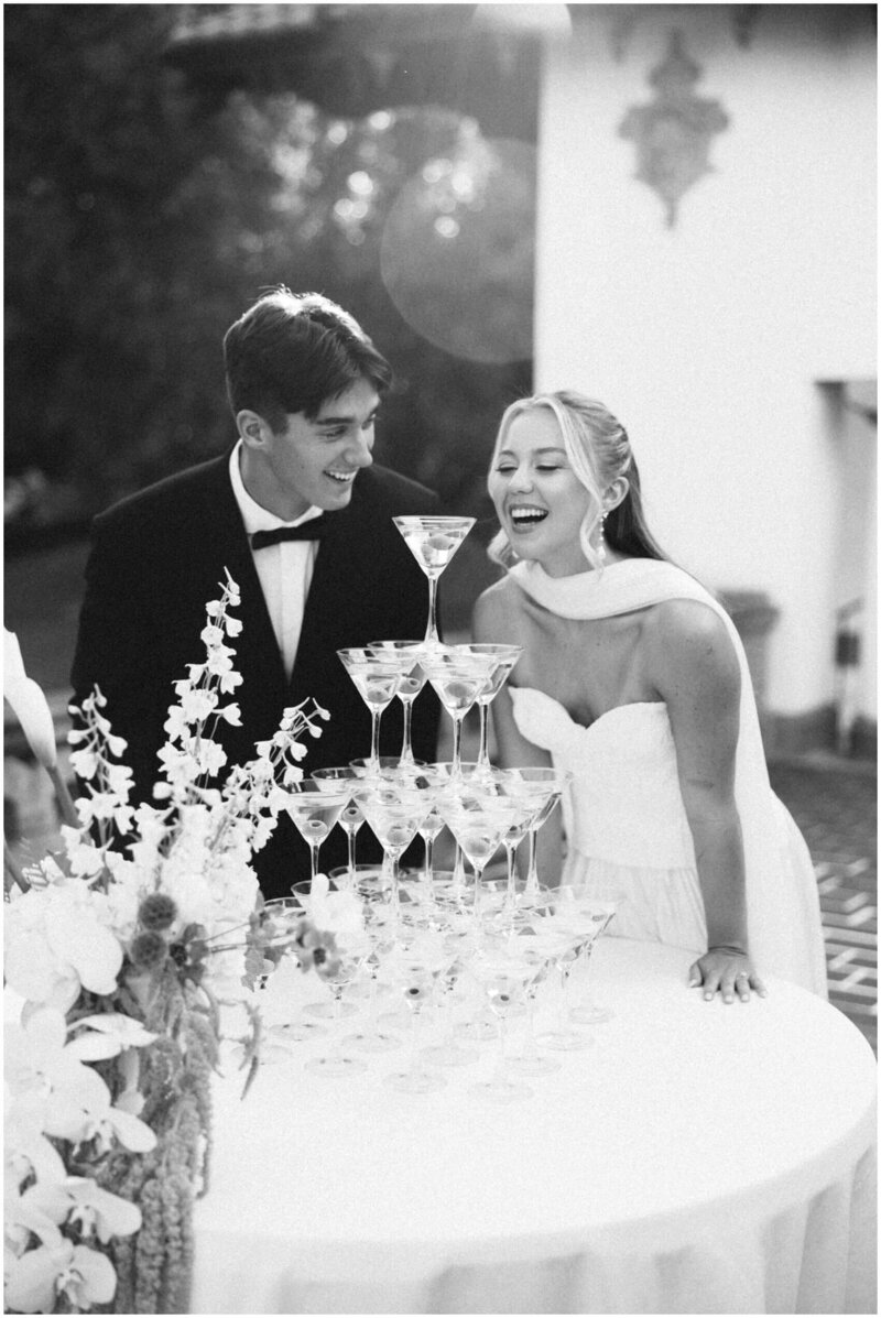 A bride and groom laugh while admiring a martini tower at their wedding captured in black and white by a film wedding photographer