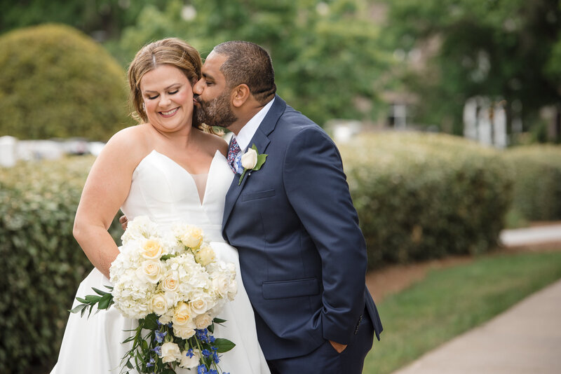Romantic wedding photography at Le Méridien Charlotte. A groom in a navy blue suit kisses his bride in a white gown holding a cascading bouquet of white roses, hydrangeas, and blue flowers. Captured by a Charlotte Indian wedding photographer specializing in timeless, emotional wedding moments.