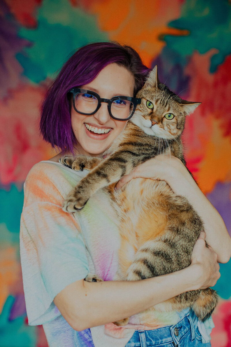 Portrait of photographer Sylvie Rosokoff holding a pixie bob cat against a rainbow backdrop by Bridget Badore