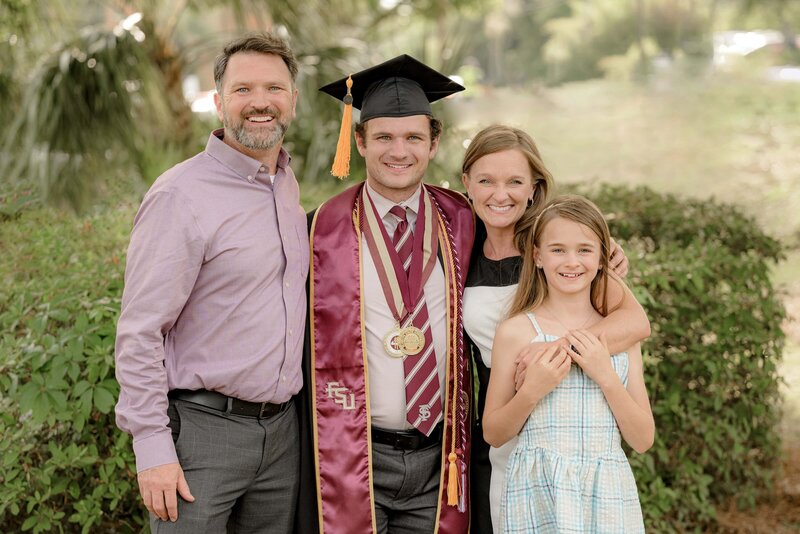 A graduate wearing a cap and gown stands with three family members outdoors, all smiling for the photo.