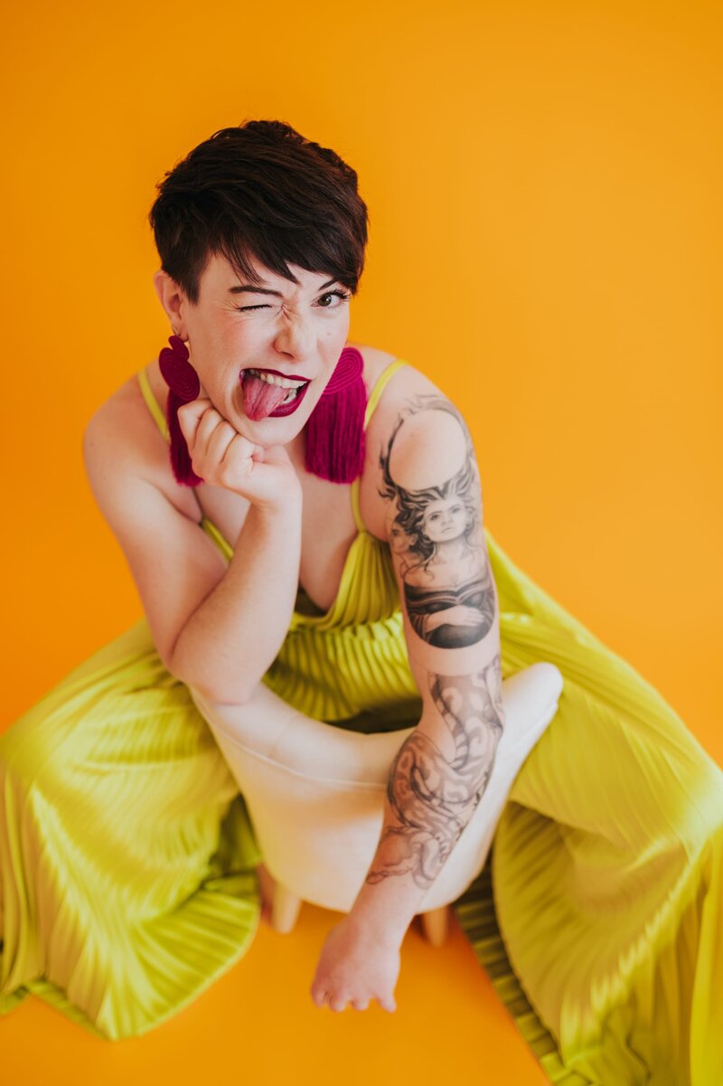 Woman with very short brown hair sits in a yellow chair wearing a bright yellow jumpsuit and long, red, fringe earrings sticks out her tongue to the camera to pose in front of yellow-orange backdrop.