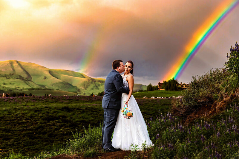Shannon Lee, a Colorado elopement photographer, captured a bride and groom embracing in a lush green field at sunset, framed by two vibrant rainbows in the sky. The scene conveys a romantic and magical tone, celebrating their love and the beauty of their elopement.