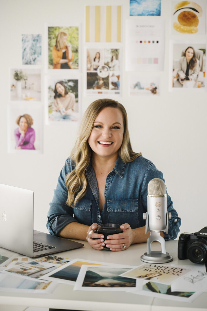 Krista Marie, a brand photographer, holding a cup of coffee and sitting at her desk with a podcast mic and brand photos on display