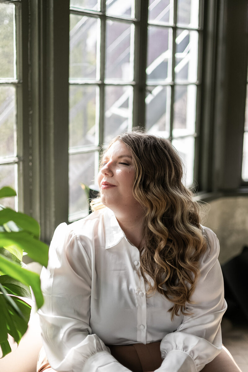 Portrait of Ashleigh Henry, multi-faceted female founder and speaker, in a white button down near plants and olive green painted windows, representing integrated ambition and lived-in wholeness.