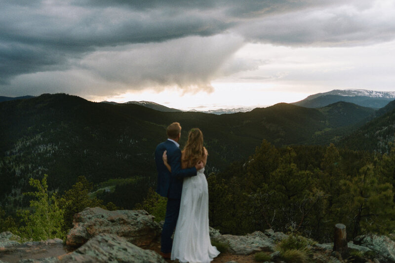 A wedding couple standing on a rocky overlook looking out over the mountains.
