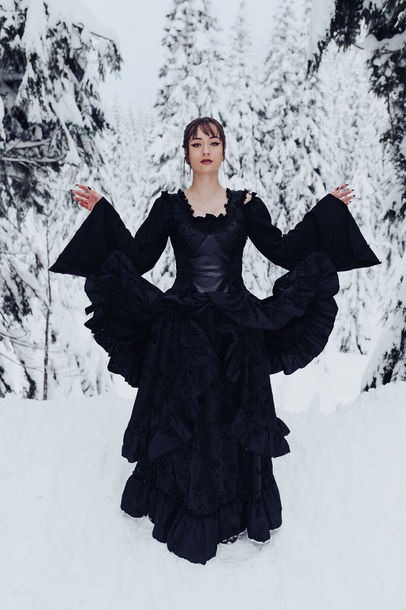 Woman stands outdoors wearing a black, gothic, floor-length gown with bell sleeves in snow-covered forest.