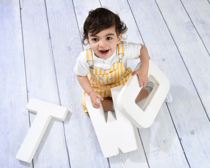 A joyful toddler in yellow striped overalls holding large "TWO" letters during a vibrant two-year-old photo shoot on a white wooden floor.