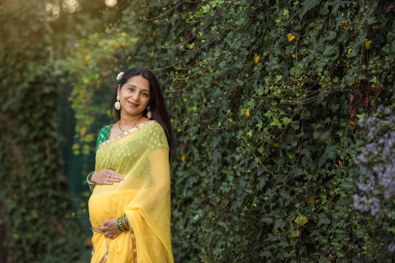 A pregnant woman holding her belly smiling next to some beautiful green vines.