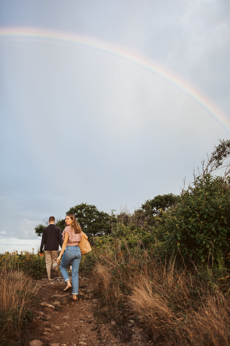 Couple walking along a coastal trail under a rainbow during their Maine engagement session.