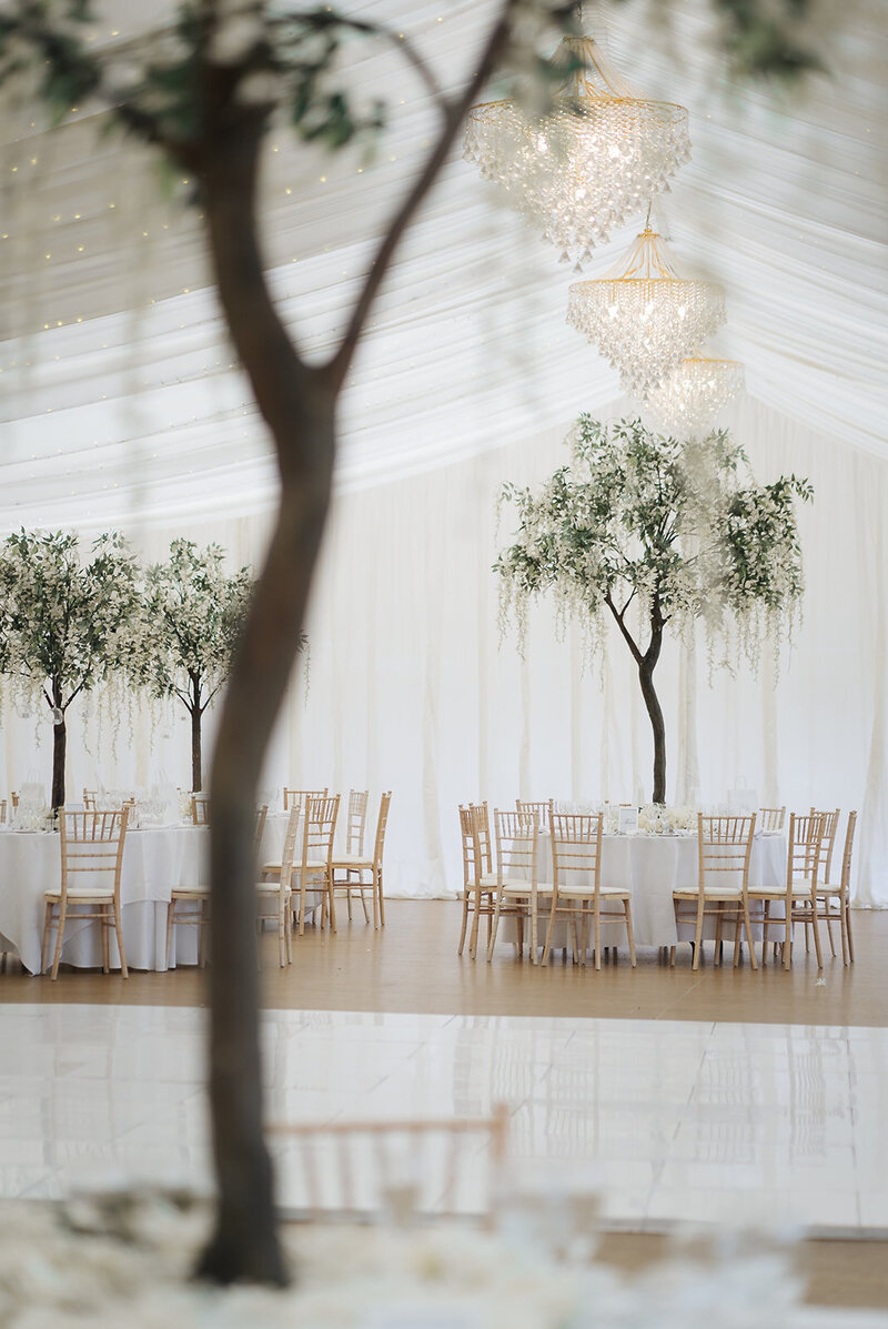 A marquee wedding set up with indoor trees, round white tables and a white dance floor.