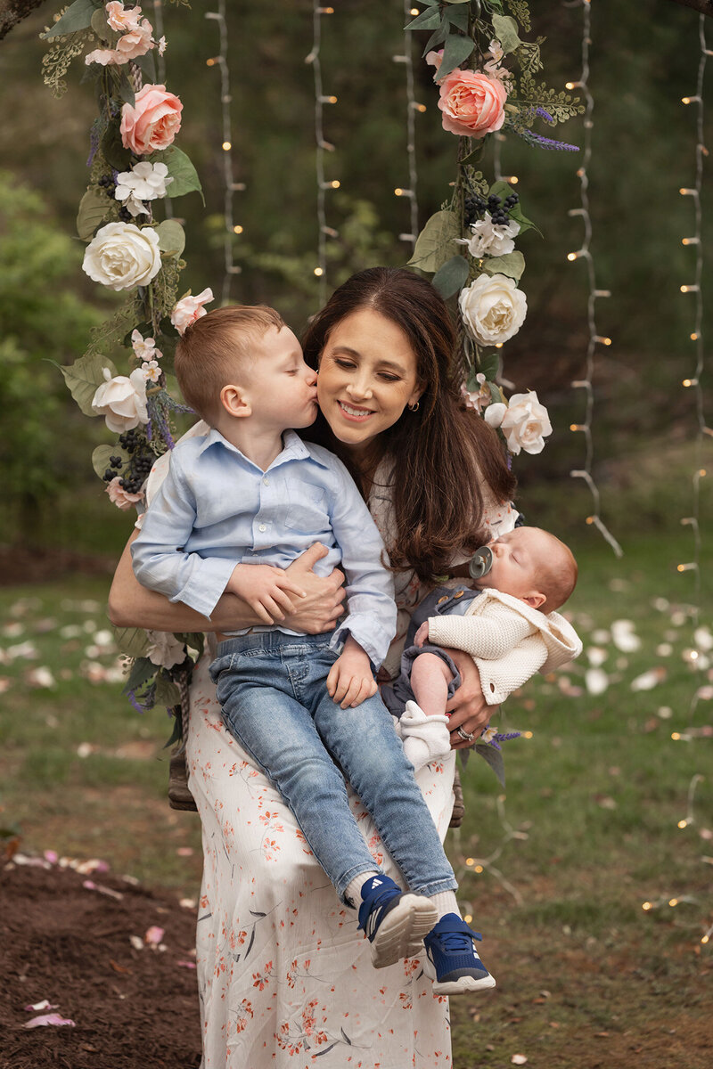 Mom sits on swing with her two songs. Her older son gives her a kiss while the baby sleeps in his mom's arms.