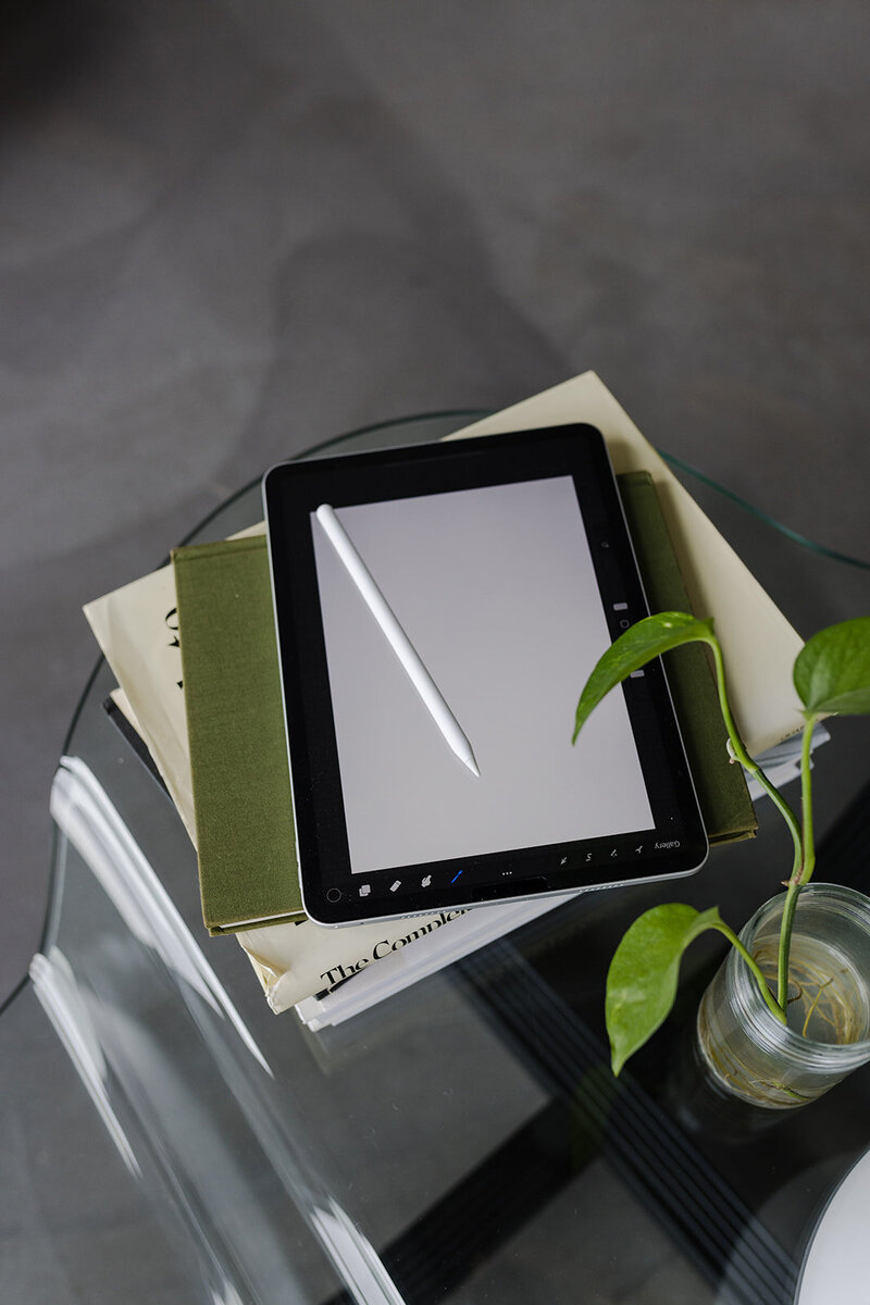 ipad and apple pencil rested on a stack of books on a glass table with concrete flooring showing underneath, a plant sits off to the side
