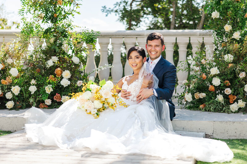 A joyful couple poses on their wedding day, surrounded by lush floral arrangements and a classic stone balustrade. The bride's elegant lace gown and the groom's sharp suit create a timeless and romantic scene under a clear blue sky.