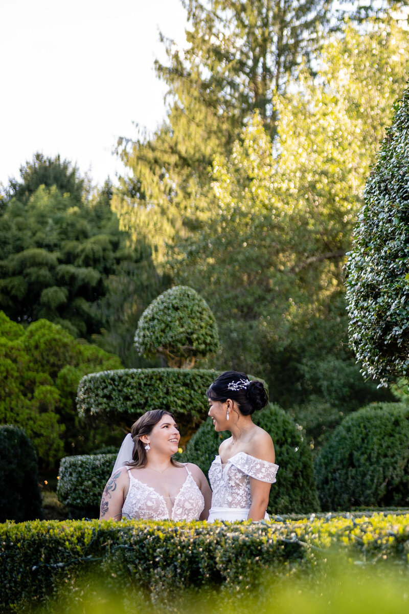 laotian bride in off the shoulder white gown with a curvy white bride in a spaghetti-strap gown smiling at each in green hedge garden