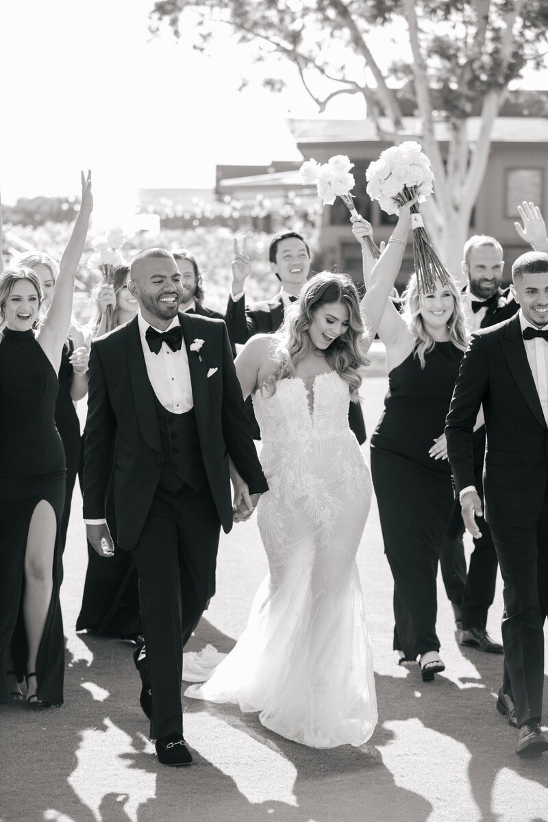 A bride and groom walking with their wedding party as the bride holds up her bouquet in celebration