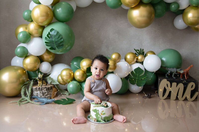 1 year old boy wearing a grey onsie with his hand in his dino cake for his First Birthday cake smash photography session in Burlington, Ontario.