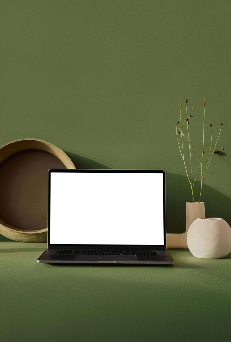 A sleek laptop with a blank white screen is set on a modern green surface, accompanied by minimalist ceramic vases and a neutral-toned decorative bowl against a matching green backdrop. This contemporary setup represents the perfect balance of productivity and relaxation, inspired by vacation fundraising to support meaningful experiences.