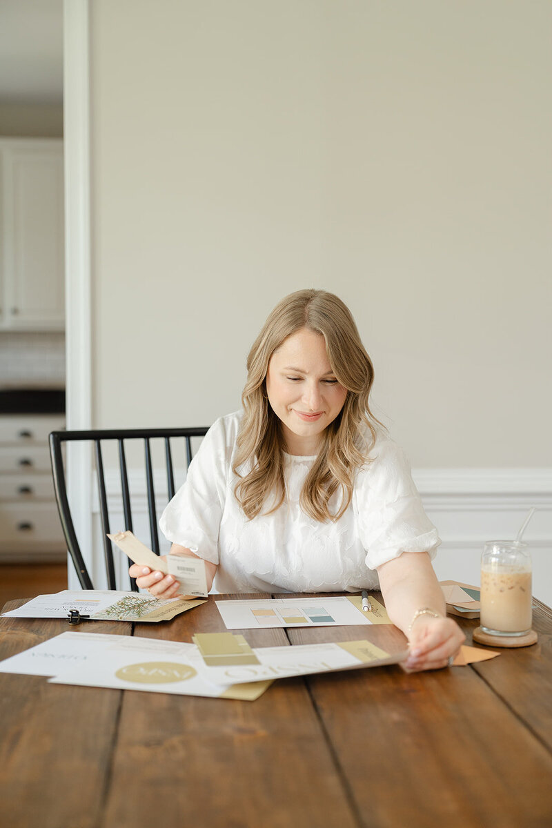 Woman sitting at a wooden table smiling and assessing various brand elements on paper