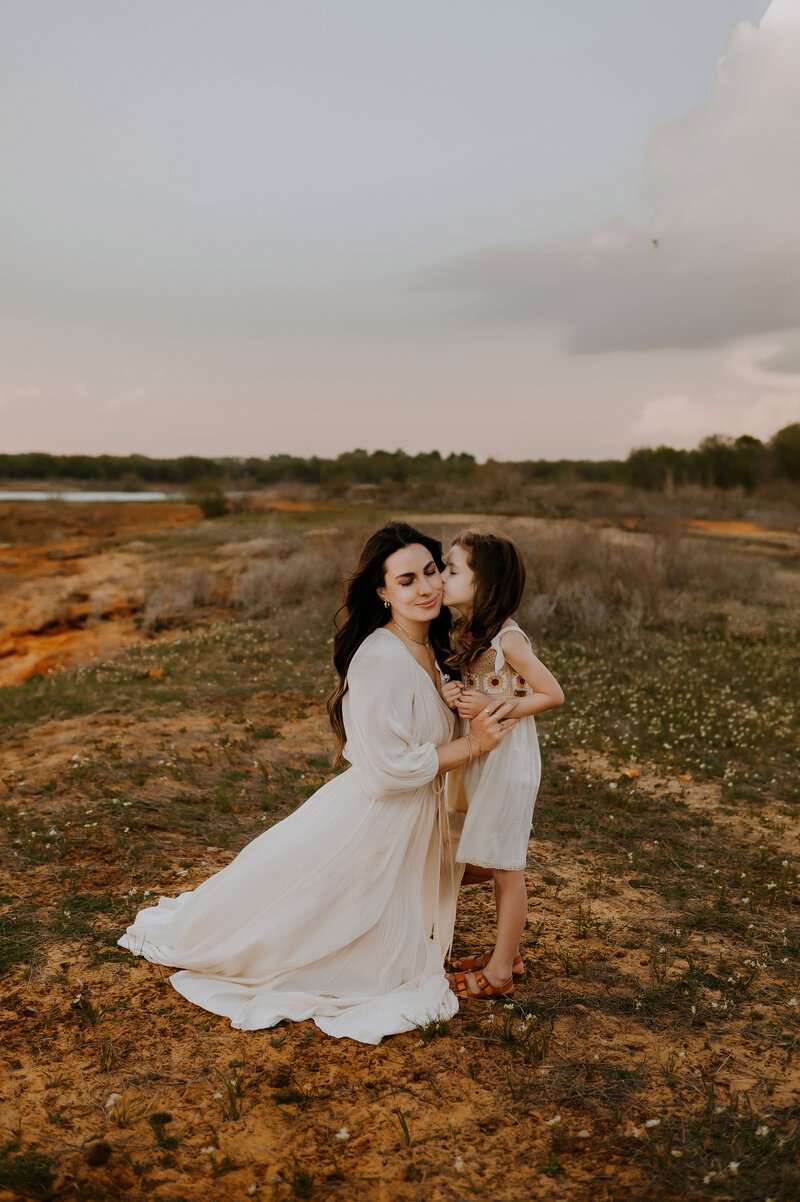 A mom is kneeling down next to her daughter while she leans in and kisses the mom on her cheek at Murrell Park in Flower Mound, Texas