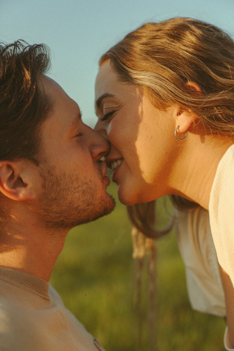 Close-up of a smiling couple sharing a playful and intimate nose kiss during golden hour in a grassy field, capturing warm light and romantic connection.