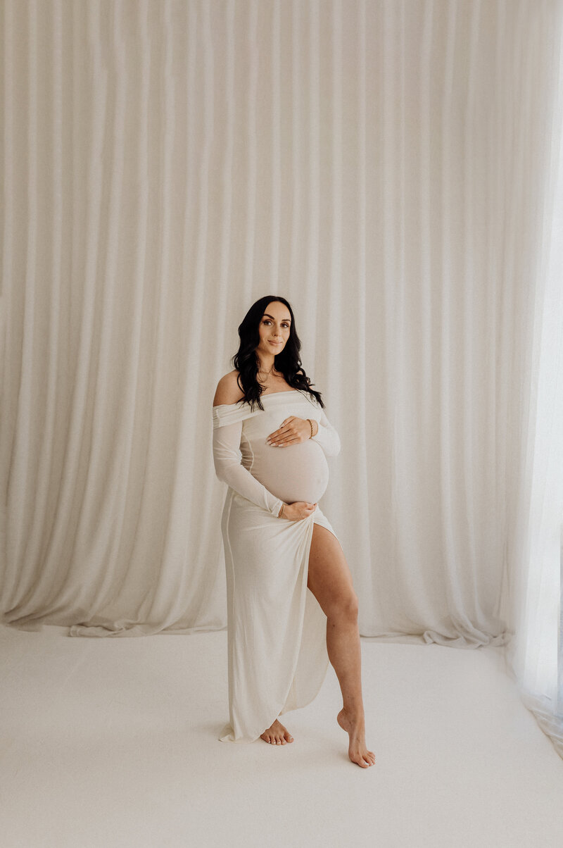 A mother to be wears a white dress with a split from lucy janes photography client wardrobe, a professional maternity photographer in christchurch