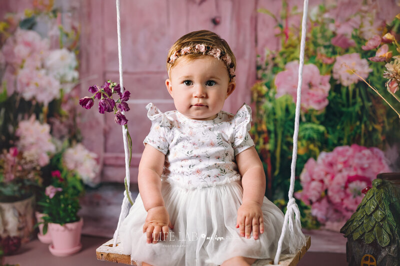 pretty toddler girl sitting on a swing infront of a pink toned  beautiful flower backdrop with a blue rustic door. 