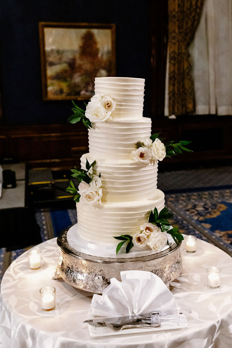 White 4-tiered wedding cake with a few white roses on each tier with greenery sits on ornate silver platter atop table with silk patterned table cloth.