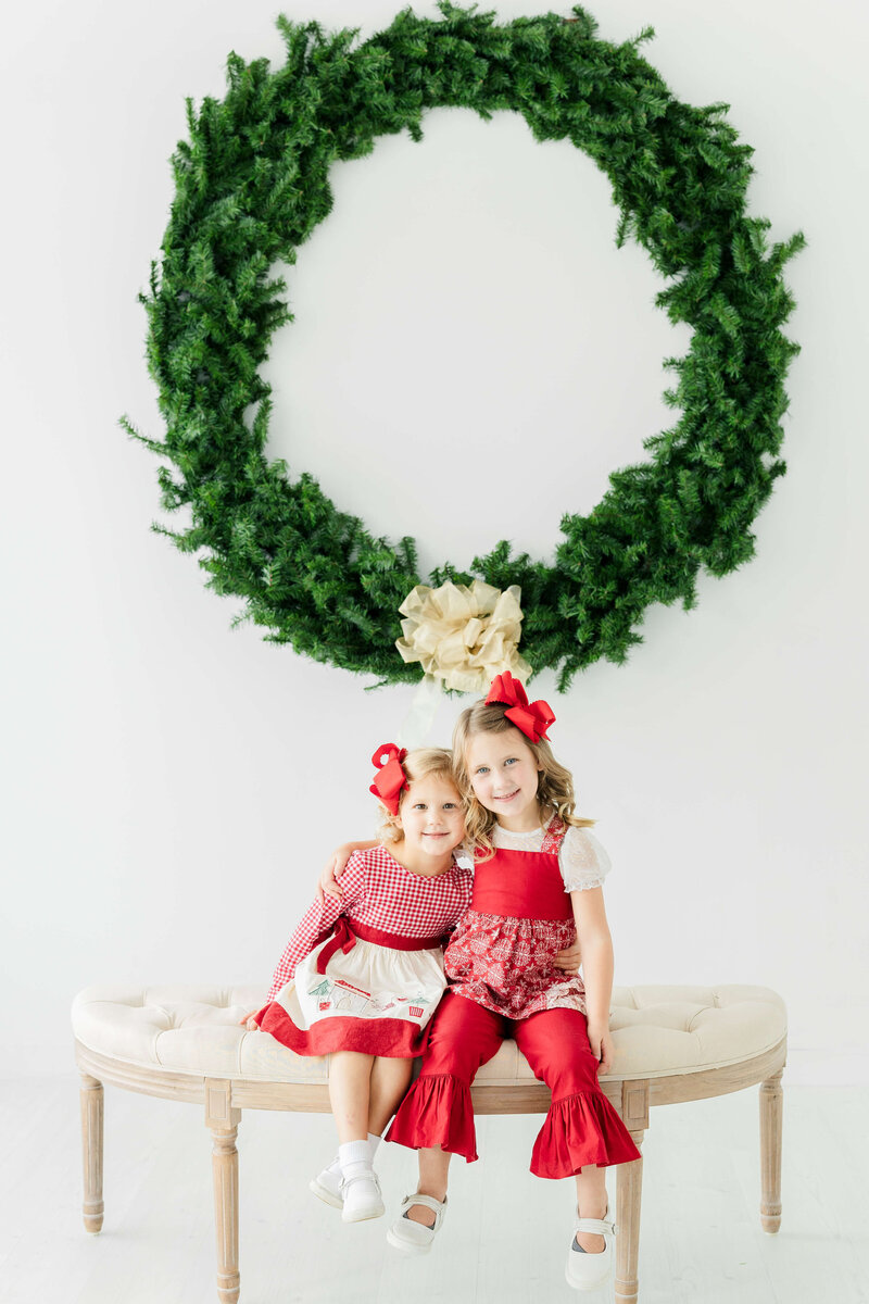 Sisters sitting on bench in front of a wreath for christmas mini sessions in Dothan Alabama