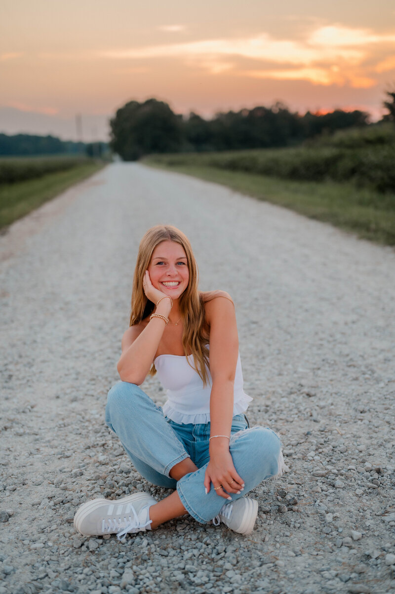 Senior girl sits in the middle of the gravel road with her chin resting in her hand while smiling at the camera.