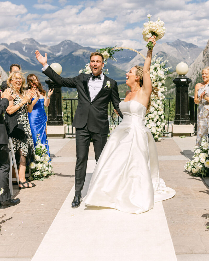 A joyful moment as the bride and groom raise their arms in celebration while walking down the aisle at the Banff Springs Hotel terrace, surrounded by clapping guests on a beautiful summer day.