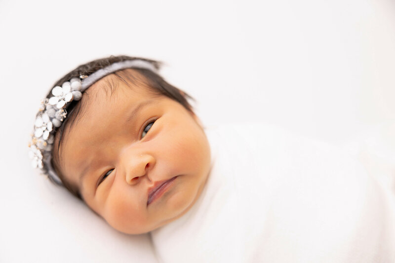 Baby girl posed for newborn photo session wrapped in white and wearing flowered headband. 